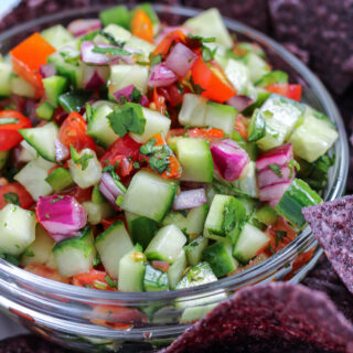 a glass bowl of cucumber pico de gallo/ cucumber salsa surrounded by blue corn tortilla chips.